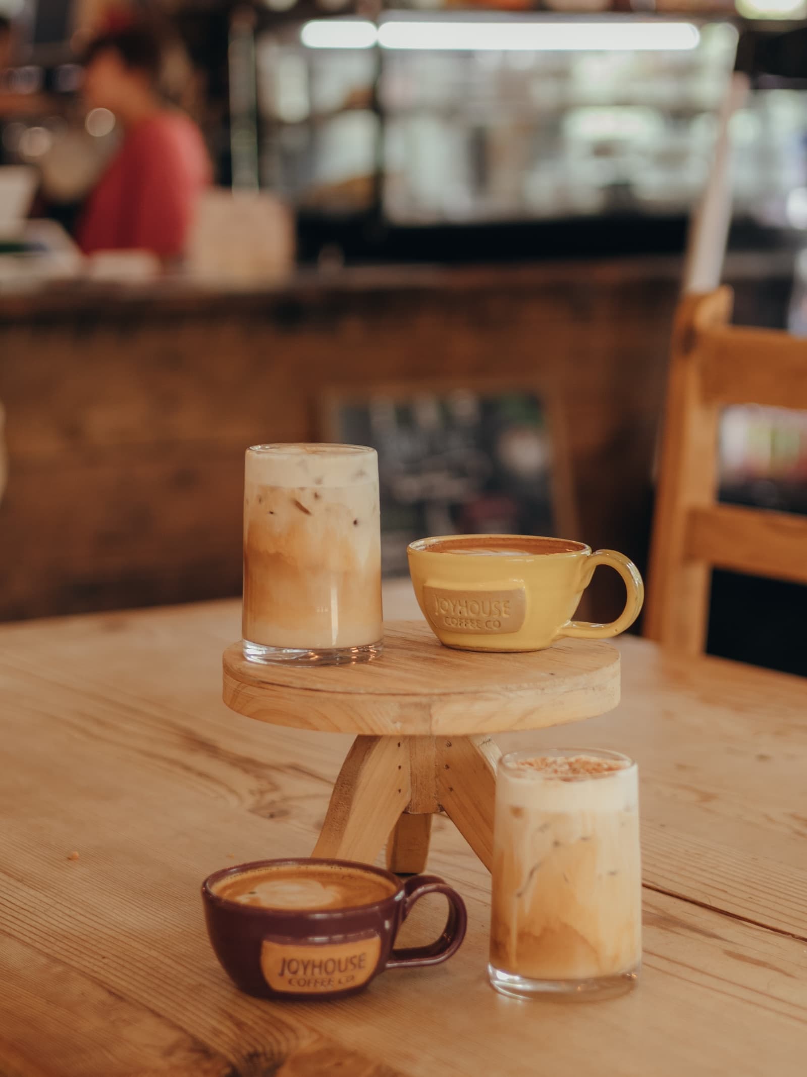 Lattes in Joyhouse mugs and two iced lattes on a cafe table, the bar and a barista behind